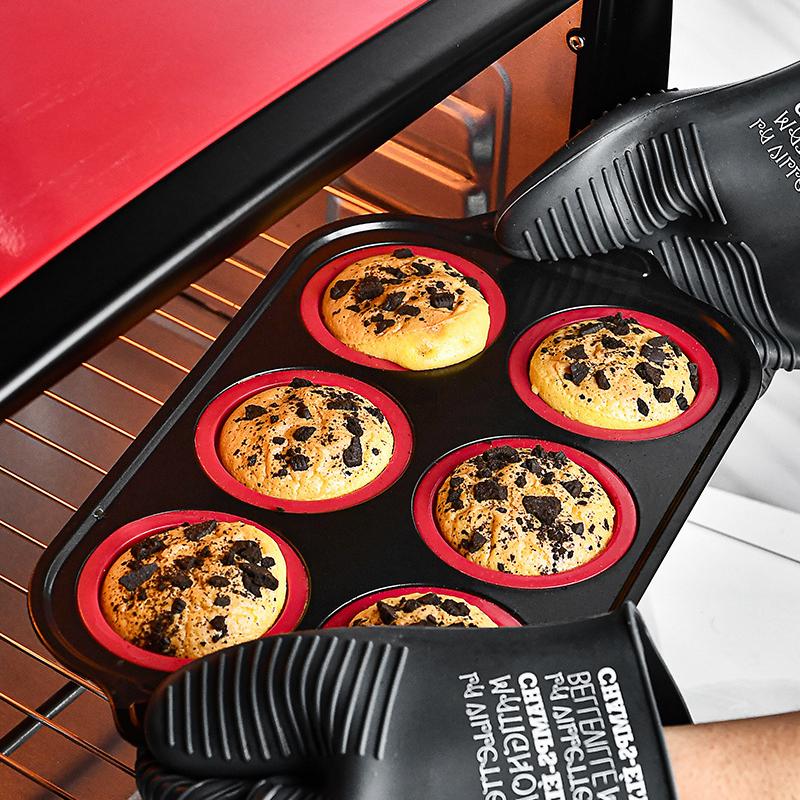 Red six-cavity silicone baking mold filled with chocolate chip cakes and held with oven mitts in front of an open oven
